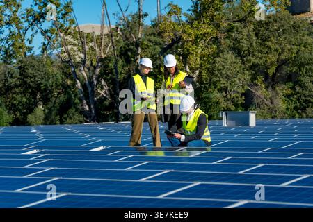 Diverse group of engineers wearing hard hats and safety vests using clipboard, laptop, and tablet while inspecting large solar panel array on build... Stock Photo