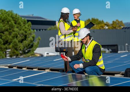 Diverse group of engineers wearing hard hats and safety vests using clipboard, laptop, and tablet while inspecting large solar panel array on build... Stock Photo