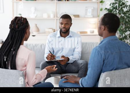Black male psychologist having therapy session with couple and taking notes while sitting on couch in office Stock Photo