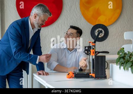Asian male industrial engineer wearing eyeglasses sitting at table and presenting 3D printer to caucasian male colleague standing beside him in office Stock Photo