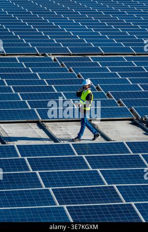 Hispanic male engineer in hard hat and safety vest talking on mobile phone and holding tablet while walking on commercial rooftop with solar panels... Stock Photo
