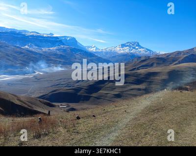 Snow-capped mountains rise above valley with dry grasses. Smoke rises from distant hills under clear blue sky. No animals or plants identified in imag Stock Photo