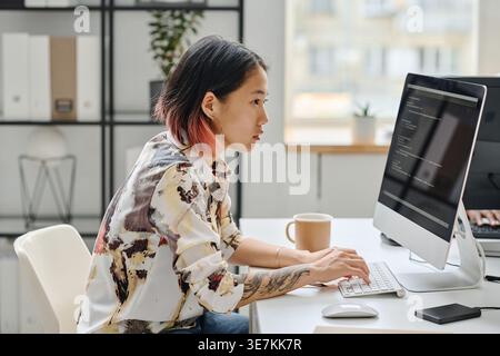 Asian female programmer working on computer typing codes while sitting at desk with coffee in office Stock Photo