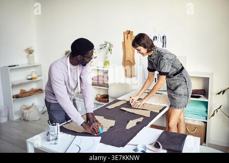 Black male fashion designer wearing eyeglasses working with caucasian female tailor standing at table in atelier Stock Photo