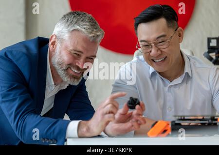 Asian and caucasian male industrial engineers examining product created by 3D printer while sitting at table working together in office Stock Photo