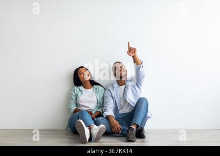 Dreaming african american couple planning interior, decorations and furniture in their own house, sitting on floor and looking up, man pointing at ... Stock Photo