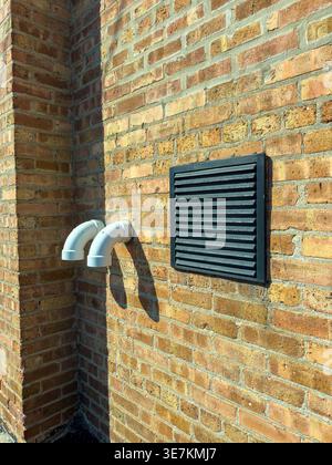 White pipes extend from the wall next to a black vent. The wall is made of bricks. Bright sunlight shines on the structure. The setting is in a city a Stock Photo