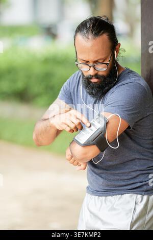 Mixed-race man with earphones using smartphone on armband standing in park during daytime Stock Photo