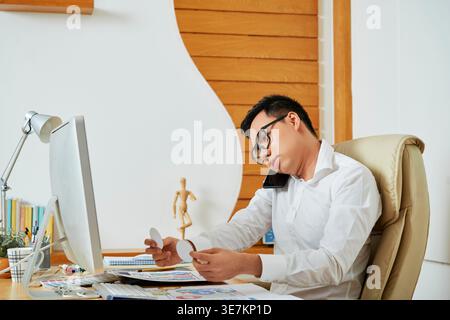 Asian male graphic designer having phone call using smartphone while rearranging icons sitting on chair in office Stock Photo