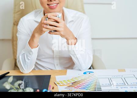 Cropped image of Asian male graphic designer holding cup of coffee sitting at table in office Stock Photo