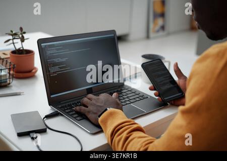 Black man holding smartphone working on laptop sitting at table in office Stock Photo