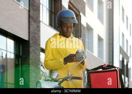 Black delivery man wearing helmet using smLifestylephone standing near motorcycle with food delivery box on city street during daytime Stock Photo