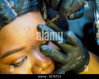Beautician wearing black gloves is carefully applying permanent makeup on a customer's eyebrows using a tattoo machine Stock Photo