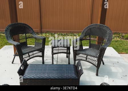 Two black wicker chairs and a small table are arranged on a concrete surface in a garden space surrounded by a wooden fence and green grass under clea Stock Photo