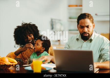 Serious Black man working on laptop sitting at table with his wife and son eating breakfast at home Stock Photo