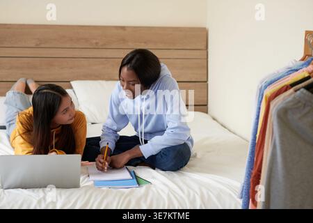 Smiling Black female student sitting on bed with crossed legs with pencil and notebook on hand discussing with Asian female friend inside a room Stock Photo