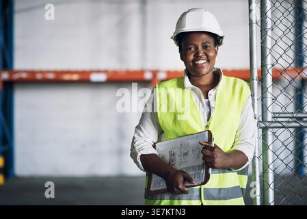 Happy woman, portrait or civil engineer with clipboard in warehouse for industrial construction. Female person, contractor or builder with floor plan Stock Photo