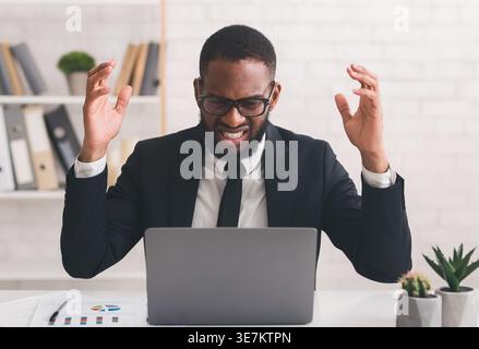 Black businessman with angry expression raising hands  looking at laptop while sitting at desk in office Stock Photo
