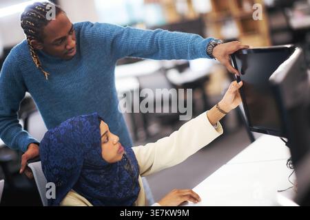 Focused Black man using computer standing behind Black muslim female coworker sitting during meeting in office Stock Photo