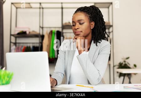 Black female fashion designer using laptop leaning chin on hand while sitting at table in fashion boutique Stock Photo
