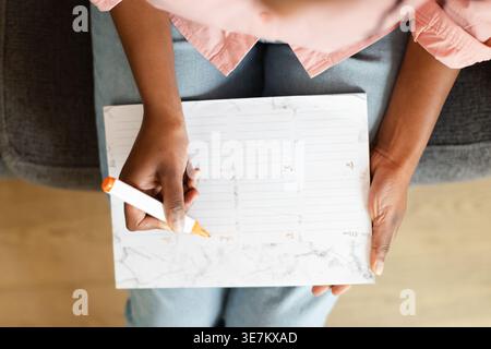 Cropped image of Black woman making her daily reminder agenda on notes while sitting on sofa in living room at home Stock Photo