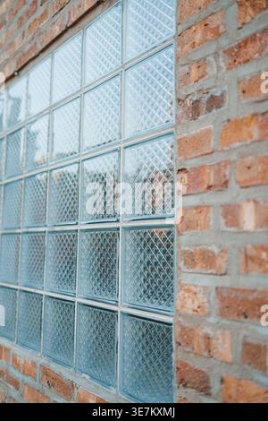 The glass block window shows patterns through clear squares. The surrounding brick wall has a rugged appearance and is exposed to sunlight. Stock Photo