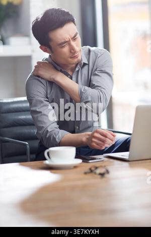 Stressed Chinese man touching shoulder with closed eyes having problem at work while sitting at table with laptop in office Stock Photo