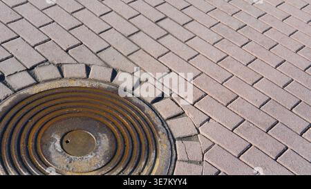 Rusty weathered manhole cover on tiled pavement with brick pattern, copy space. City infrastructure, safety in maintenance Stock Photo
