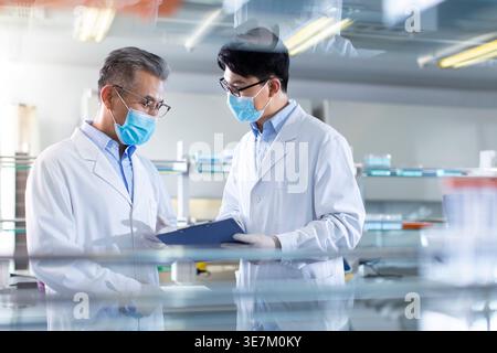 Chinese male researchers wearing lab gowns and face masks standing with clipboard discussing medical results in laboratory Stock Photo