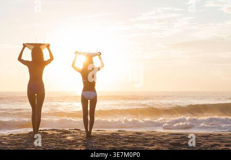 Asian and Mixed race female friends in bikinis standing with surfboards above head on beach facing sunset Stock Photo