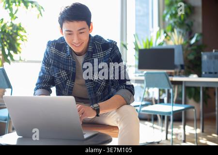 Chinese male college student leaning arms on legs sitting at table using laptop in computer room Stock Photo