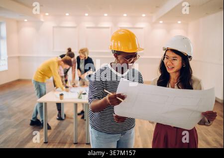 Cheerful Black women and Asian woman wearing safety helmets working on blueprint standing in office Stock Photo