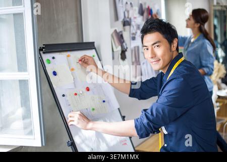 Smiling Chinese male fashion designer with measuring tape around his neck standing and working on whiteboard near female colleague in shop Stock Photo