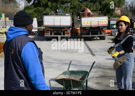 Serious Hispanic woman wearing denim overallss yellow hardhats and protective gloves holding shovel talking to Black foreman at construction site d... Stock Photo