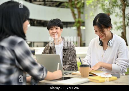 Smiling Asian male and female college students sitting at table studying together in campus park during daytime Stock Photo