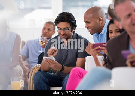 Asian businessman wearing eyeglasses using smLifestylephone with Black male colleague sitting on sofa near coworkers in office event Stock Photo