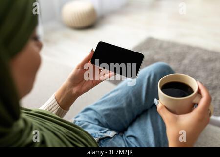 Cropped view of young Arab woman in hijab using smartphone with blank screen, sitting on sofa with coffee, mockup for mobile app or website design. Te Stock Photo