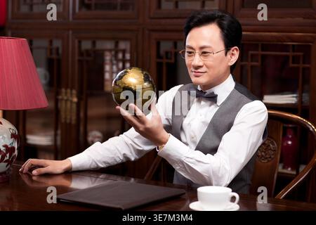 Chinese businessman holding globe sitting at table in study room at home Stock Photo