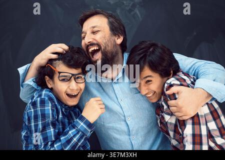 Cheerful Middle Eastern male teenagers in checkered long sleeves being hugged by caucasian man while standing against chalkboard background in studio Stock Photo