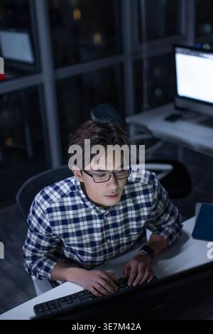 Serious Chinese man with eyeglasses wearing checkered long sleeves working late in office at night Stock Photo