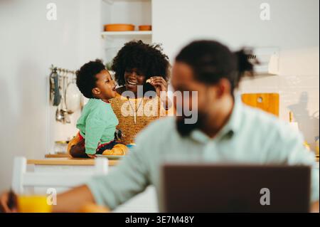 Cheerful Black mother feeding her son sitting on kitchen counter near Black father working remotely on breakfast table at home Stock Photo