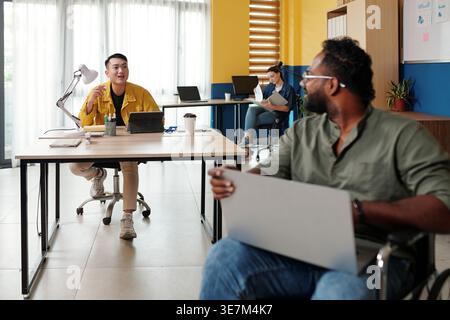 Disabled Asian man working on laptop sitting in wheelchair talking to Asian man colleague in office during daytime Stock Photo