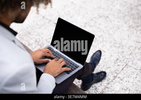 Cropped image of Hispanic businessman with curly afro wearing suit working on laptop while sitting on bench in park during daytime Stock Photo