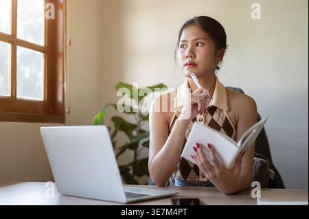 Asian woman holding pen and notebook looking away while sitting at table with laptop at home Stock Photo