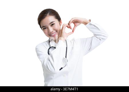 Smiling Chinese female doctor doing heart shape gesture standing against white background in studio Stock Photo