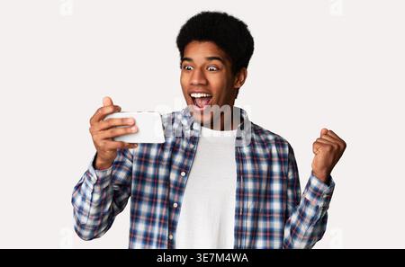 Cheerful Black male raised arm with clenched fist looking at smartphone standing against white background in studio Stock Photo