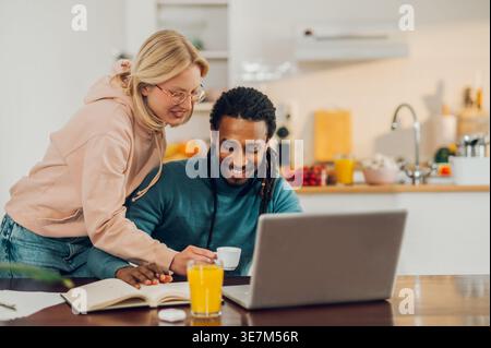 Smiling Mixed race man with locks using laptop sitting at table near caucasian girlfriend standing beside him giving cup of coffee at home Stock Photo