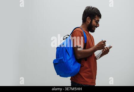 Serious Indian man in orange shirt with eyeglasses and blue backpack standing while writing down on notebook against white background in studio Stock Photo