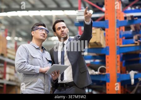 Hispanic businessman pointing finger away talking to Chinese male engineer holding tablet standing in factory Stock Photo