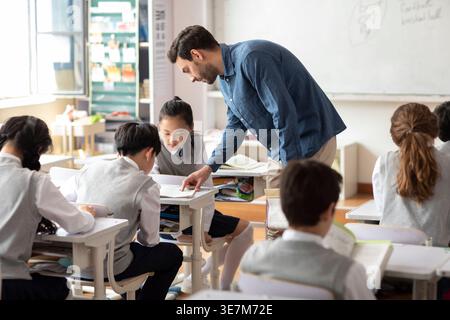 Diverse group of students doing school work while caucasian male teacher assisting them in classroom Stock Photo